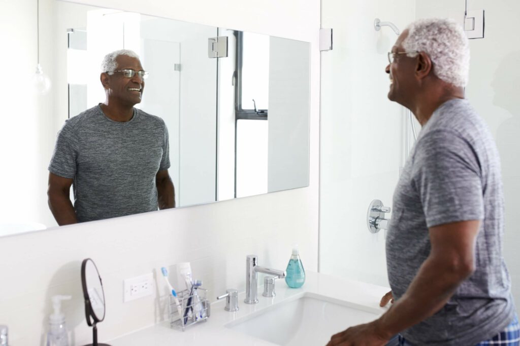An older black man smiles at himself in the bathroom mirror after brushing his teeth.
