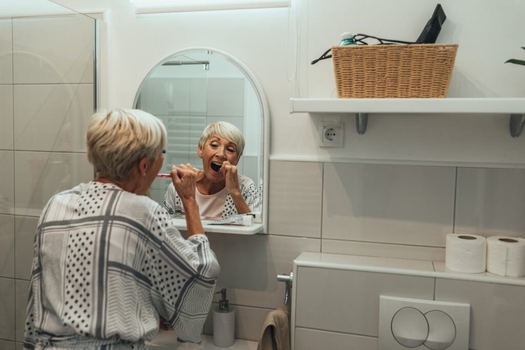 Elderly woman brushing her teeth in the bathroom mirror