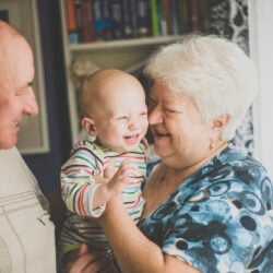 Grandparents smile and hug their baby grandson while standing in their family kitchen.