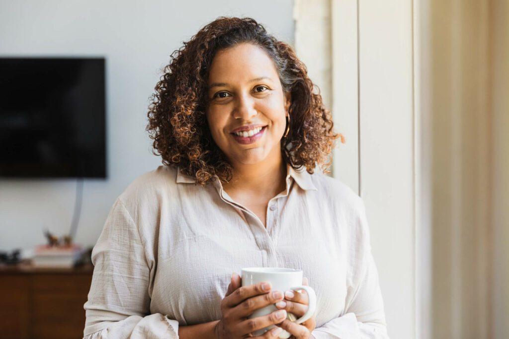 A young female carer smiles at the camera while holding a cup of team in her client's home.