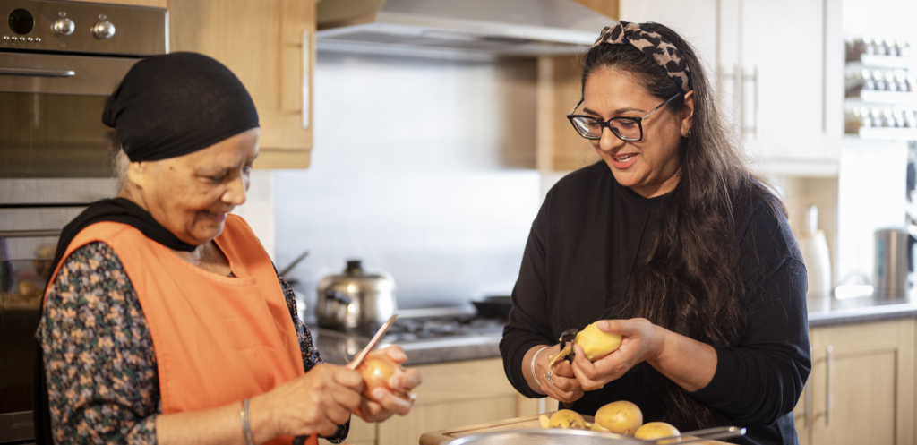 A woman helps an older lady wearing an orange apron to peel potatoes safely at home.