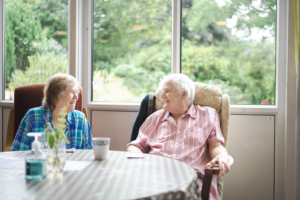An older man and woman have a chat in a sunny care home room over a cup of tea