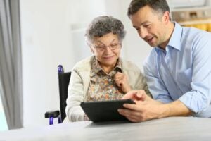 A young man shows an elderly woman in a wheelchair information on a tablet computer.