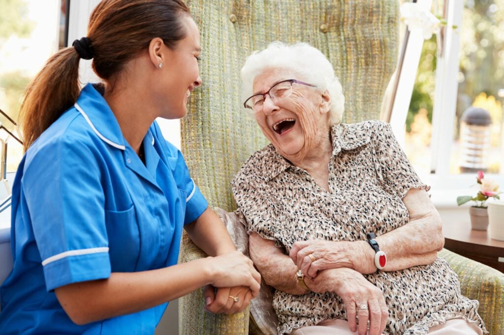 An elderly lady enjoys a laugh with a nurse wearing a uniform while sat outside on a terrace.