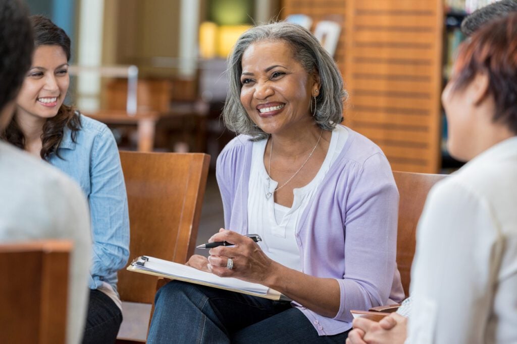 A support group smiles during a conversation, while the leader of the group holds a notebook.