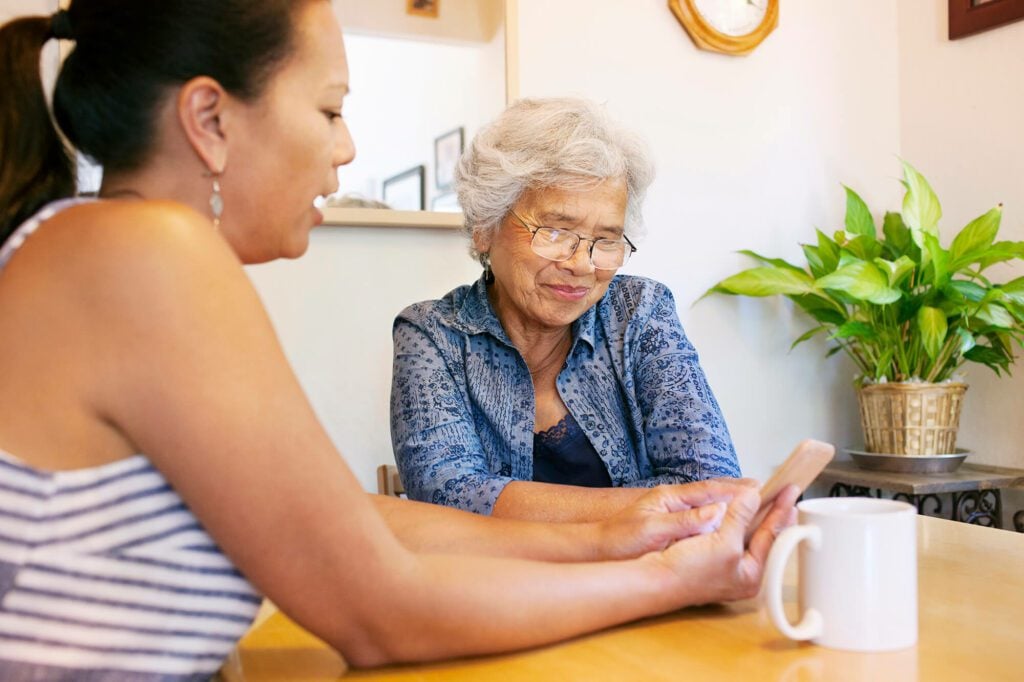 A young woman showing her elderly mother a video on her smartphone. the two are sat at a cosy breakfast table.