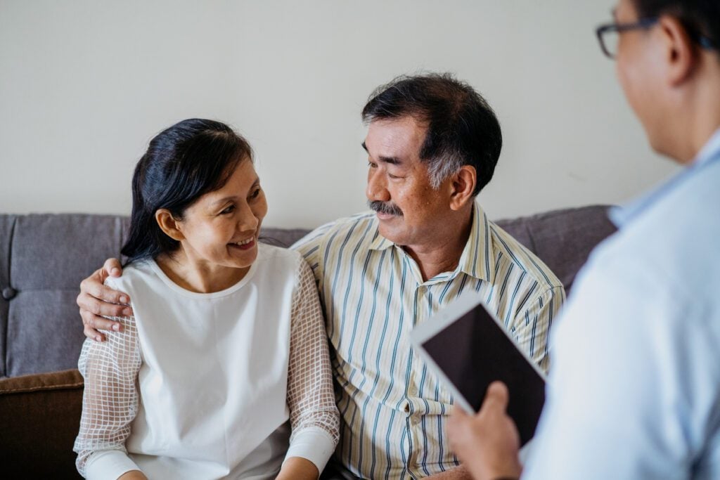 A couple smile to each other, the man with his arm round his wife as they complete an assessment with a social care professional.