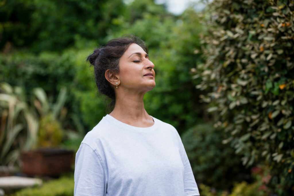 A woman wearing white stands outside in the garden taking a deep breath with her eyes closed