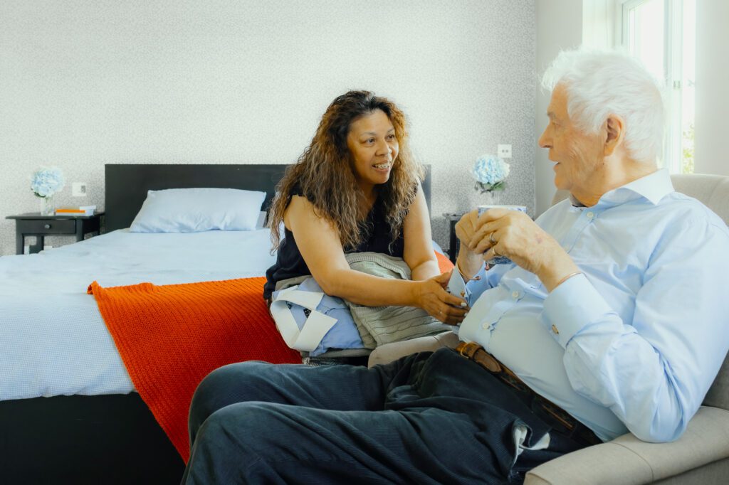 An Elder live-in carer sits on the edge of a bed while chatting to her male client over a morning cup of tea.