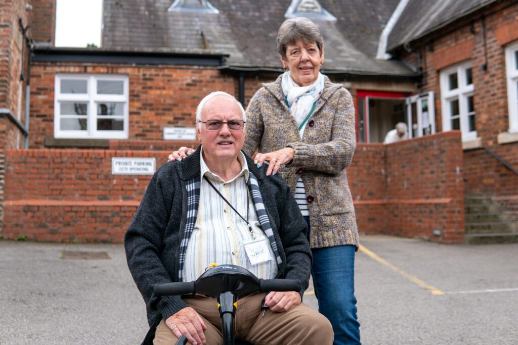 An Elderly man in a wheelchair is accompanied by his wife outside of a village community centre.