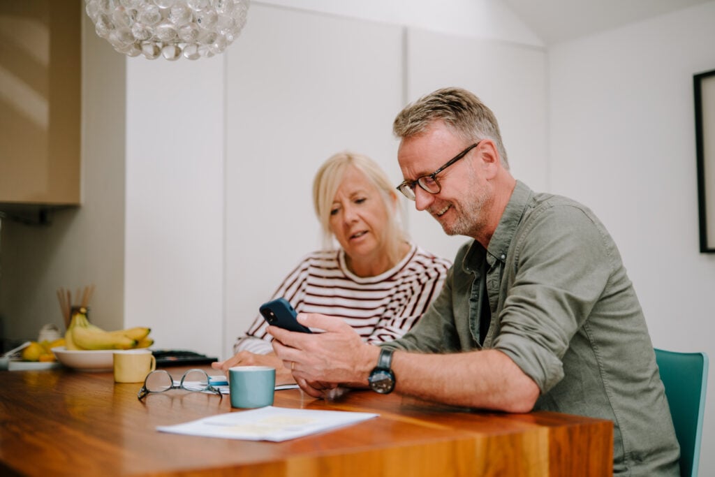 A middle aged couple research care options on a smart phone, while surrounded by financial paperwork.