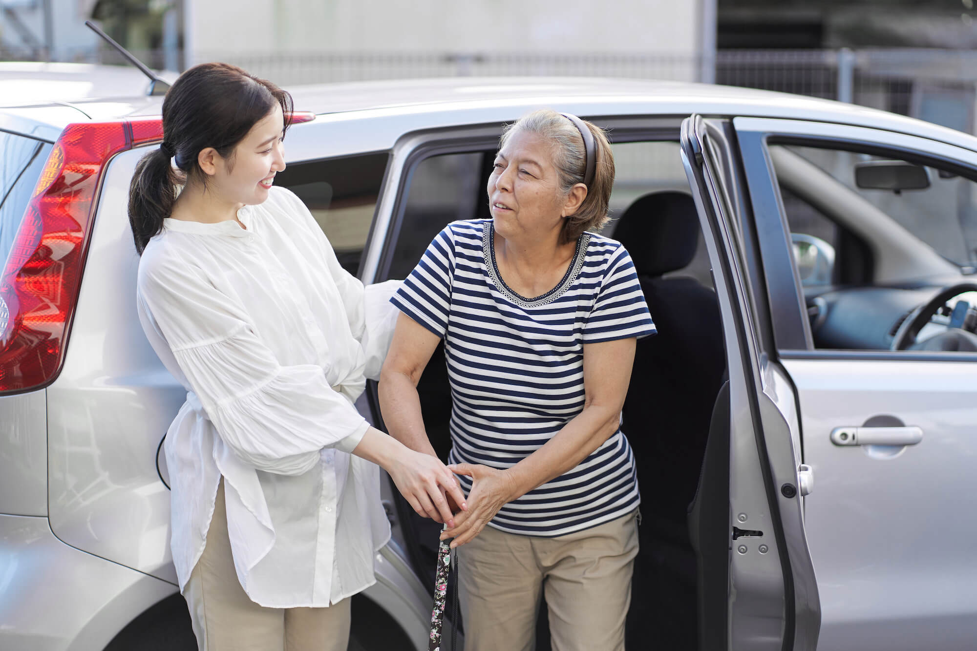 An Asian carer supporting her client as she gets out of a car with a walking stick