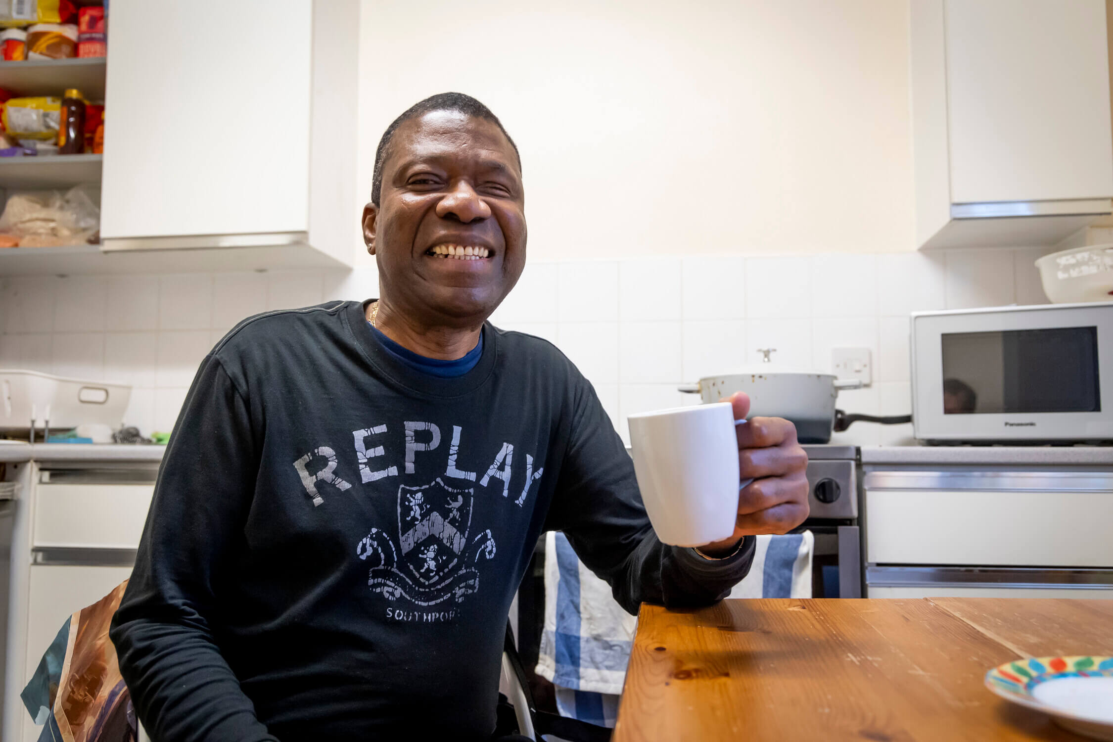 Older black man smiles while enjoying a cup of tea sat at his kitchen table.