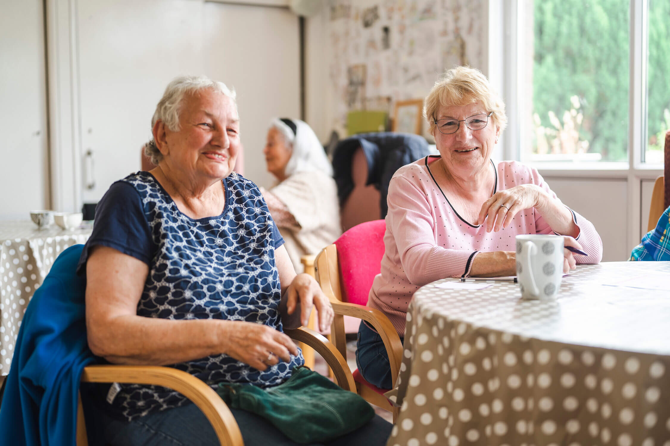two elderly ladies smiling and enjoying a cup of tea in a community dining room