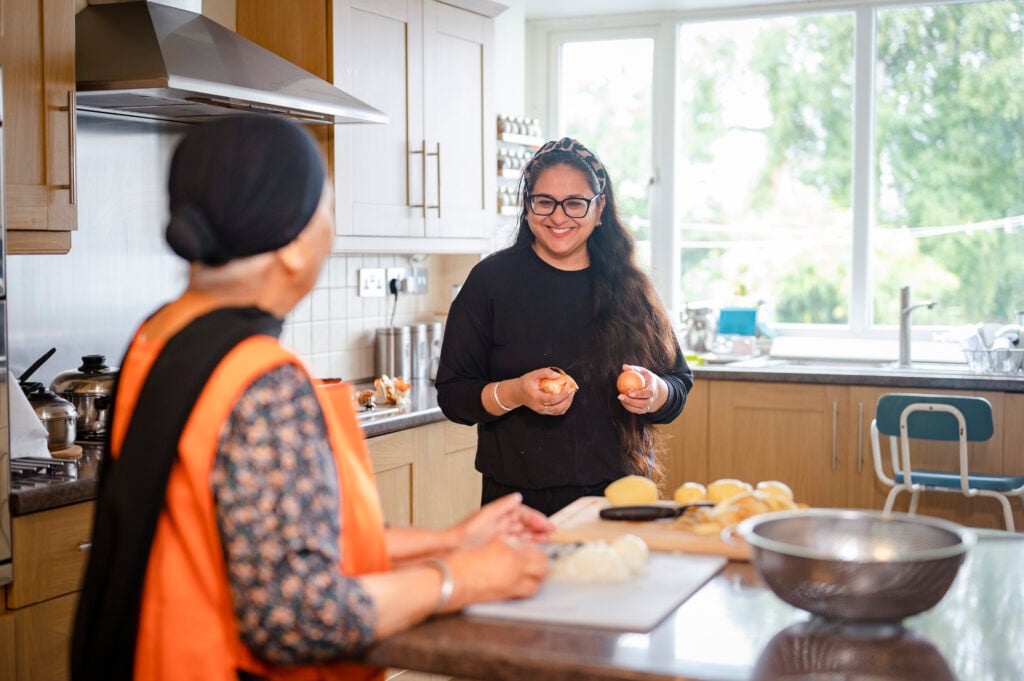 A lady helps an older woman in the kitchen to prepare potatoes.
