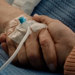 A close-up of two people holding hands, one person has a hospital IV line in their hand